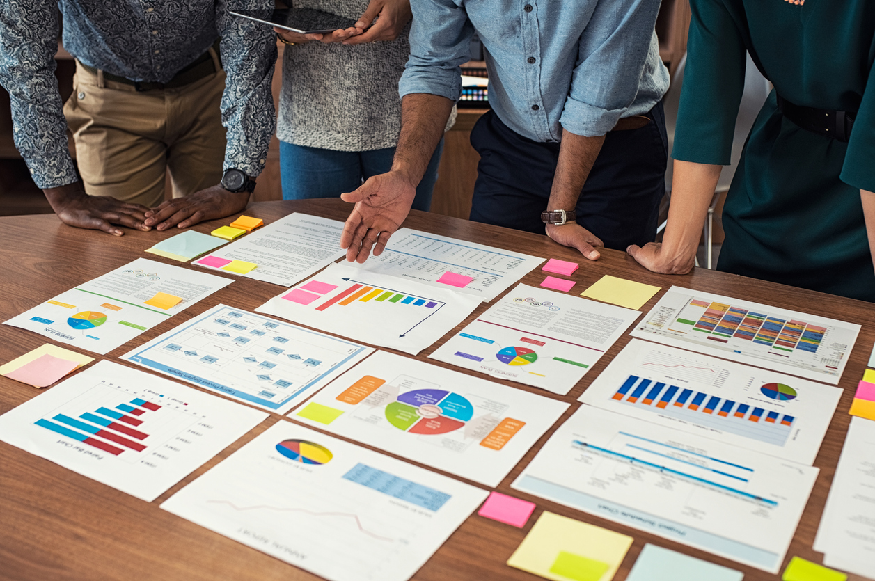 Financial and business documents on table with multiethnic hands working on it. Latin business manager with colleagues working on new startup project. Closeup business man and businesswoman hands understanding pie and bar graphs during meeting.