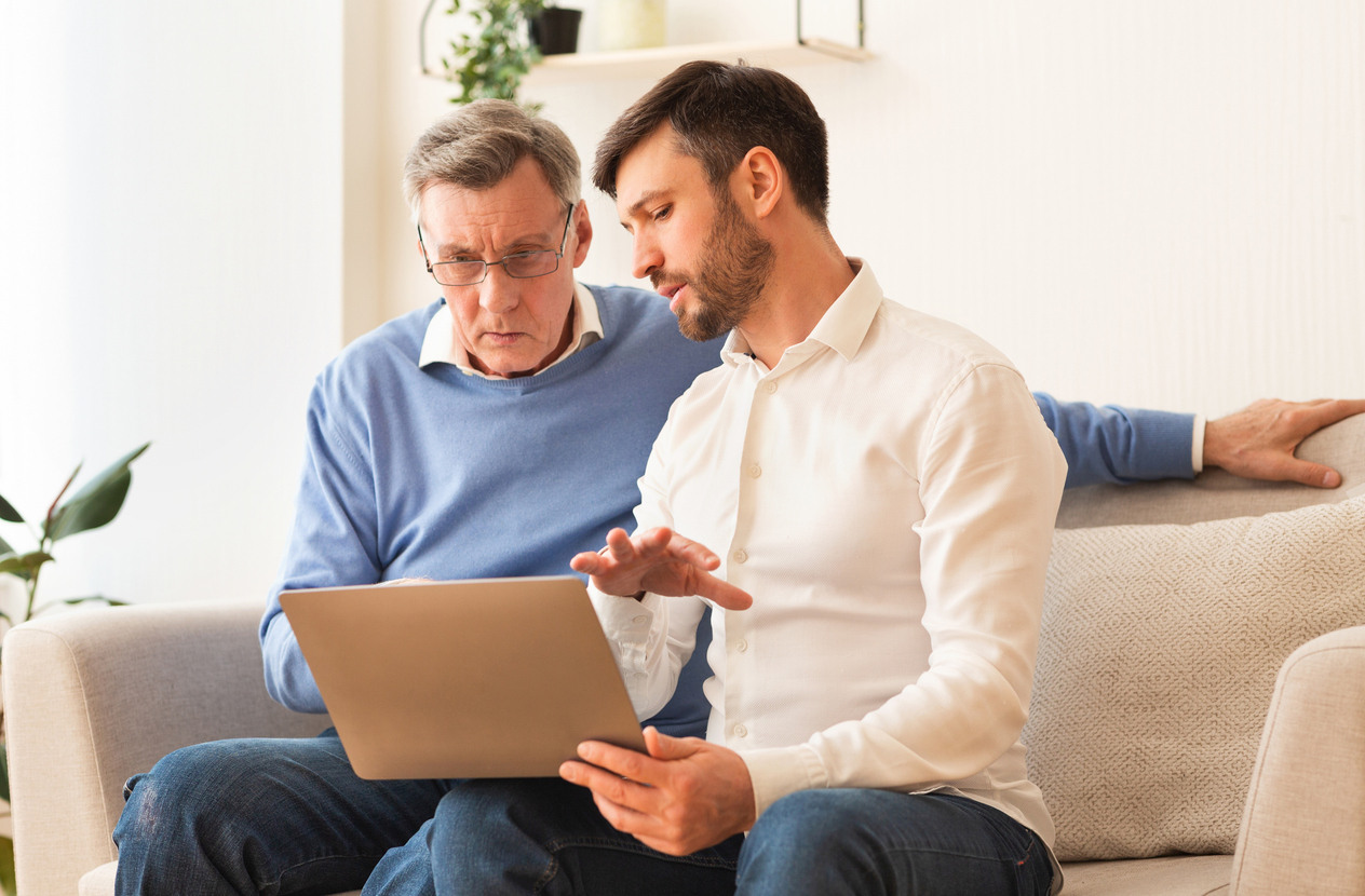 Digital Literacy. Middle-Aged Son Teaching Elderly Father To Use Laptop Computer, Social Media And Internet Sitting On Couch Indoor